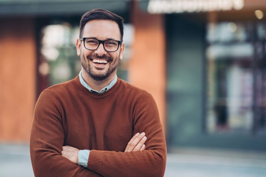 smiling man with black glasses and brown sweater standing on sidewalk
